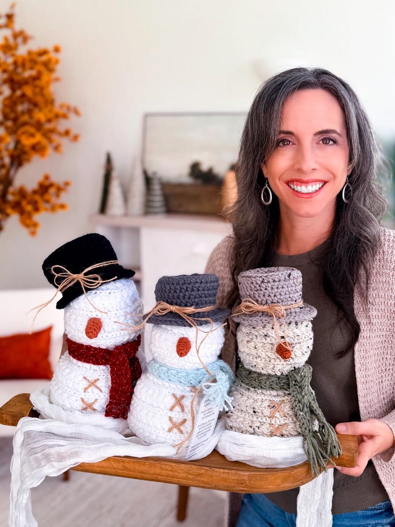 Smiling woman holding a wooden tray with three crochet snowmen in red, blue and green scarves