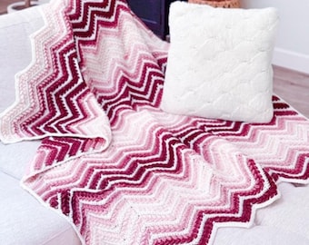 May include: A crochet blanket featuring a chevron pattern in a gradient of colours ranging from deep burgundy and dark red to light pink and white, paired with a square white textured throw pillow with a subtle embossed heart pattern.