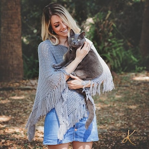 Könnte beinhalten: Eine Frau trägt einen hellblauen Strickponcho mit Fransen und hält eine graue Katze in ihren Armen. Die Frau trägt einen Jeansrock und lächelt in die Kamera.