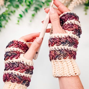 May include: Handmade crochet fingerless gloves in cream, burgundy, and purple. The gloves feature a textured stitch pattern and a cream-coloured cuff. A crochet hook is held in one hand. The gloves are designed for warmth and style.