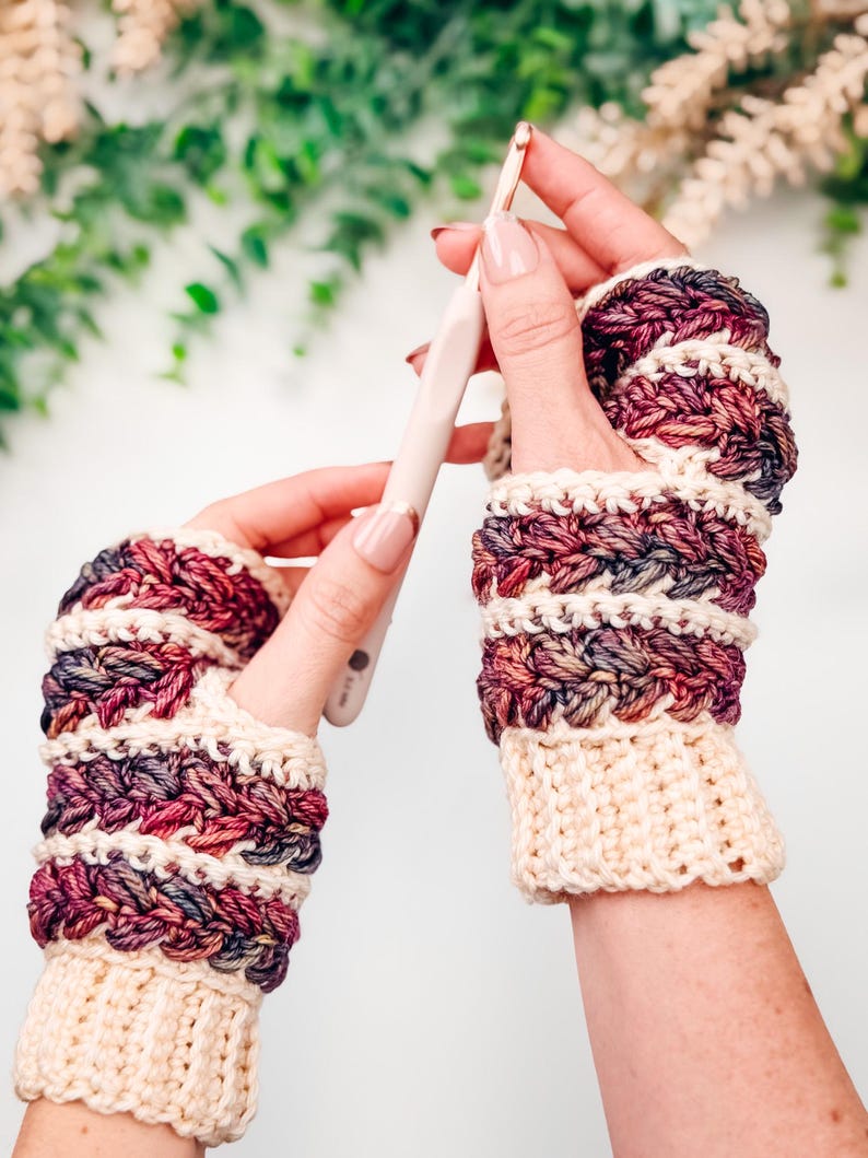 May include: Handmade crochet fingerless gloves in cream, burgundy, and purple. The gloves feature a textured stitch pattern and a cream-coloured cuff. A crochet hook is held in one hand. The gloves are designed for warmth and style.