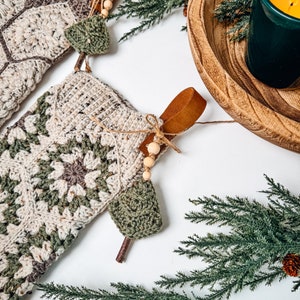 May include: Two crochet Christmas stockings featuring a cream, sage green, and brown granny square pattern. Each stocking includes a brown leather hanging loop, a small wooden bead accent, and a miniature crocheted green tree charm attached.