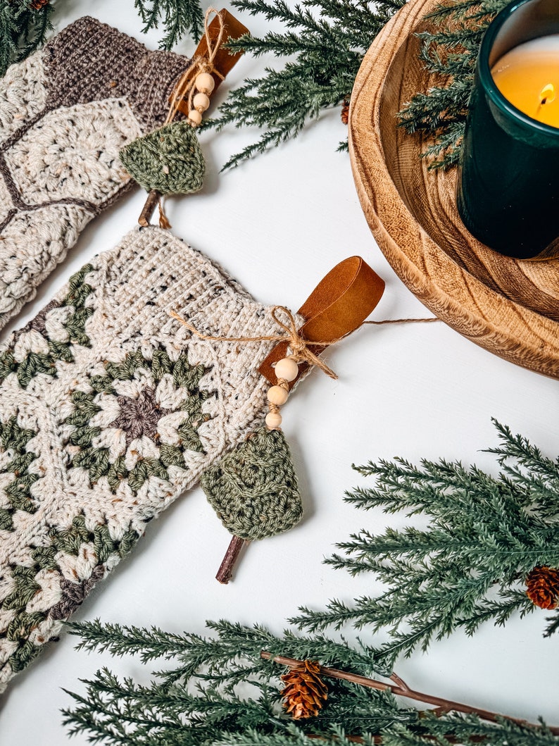 May include: Two crochet Christmas stockings featuring a cream, sage green, and brown granny square pattern. Each stocking includes a brown leather hanging loop, a small wooden bead accent, and a miniature crocheted green tree charm attached.