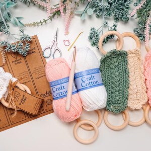 May include: A craft scene featuring crochet towel holders in green, beige, and pink, with wooden rings. Pink and white yarn balls, a crochet hook, scissors, and tags are also present. Decorative floral elements complete the composition.