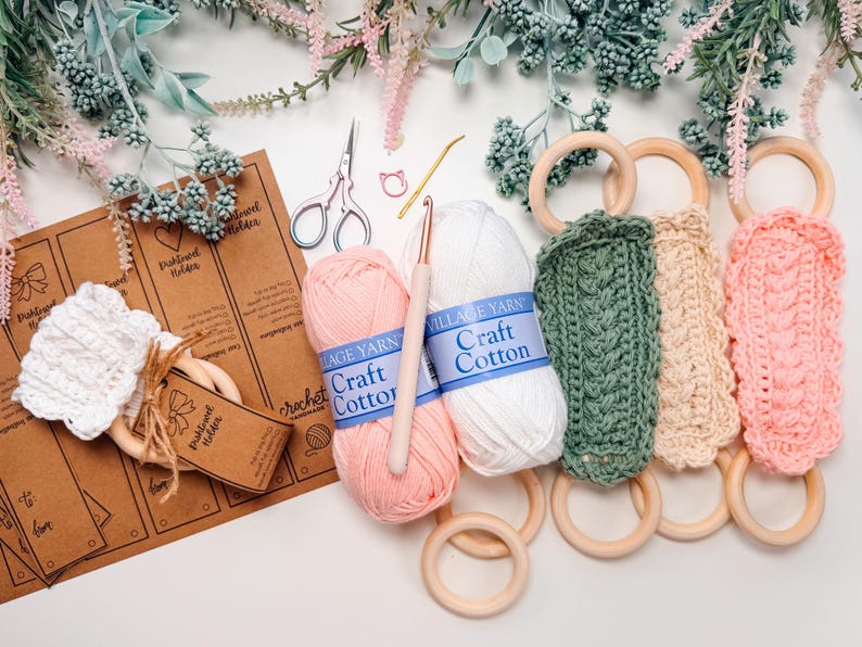 May include: A craft scene featuring crochet towel holders in green, beige, and pink, with wooden rings. Pink and white yarn balls, a crochet hook, scissors, and tags are also present. Decorative floral elements complete the composition.