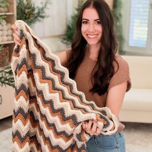 May include: A crocheted blanket featuring a repeating chevron pattern in alternating stripes of cream, tan, burnt orange, and dark charcoal gray. The textile has a textured, woven appearance with a consistent zigzag design throughout the fabric.