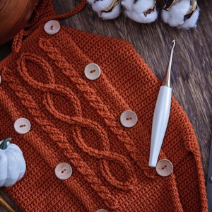 May include: A close-up of a crocheted orange baby romper with wooden buttons and a cable knit pattern. A white crochet hook is lying on the romper.