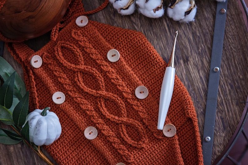 May include: A close-up of a crocheted orange baby romper with wooden buttons and a cable knit pattern. A white crochet hook is lying on the romper.