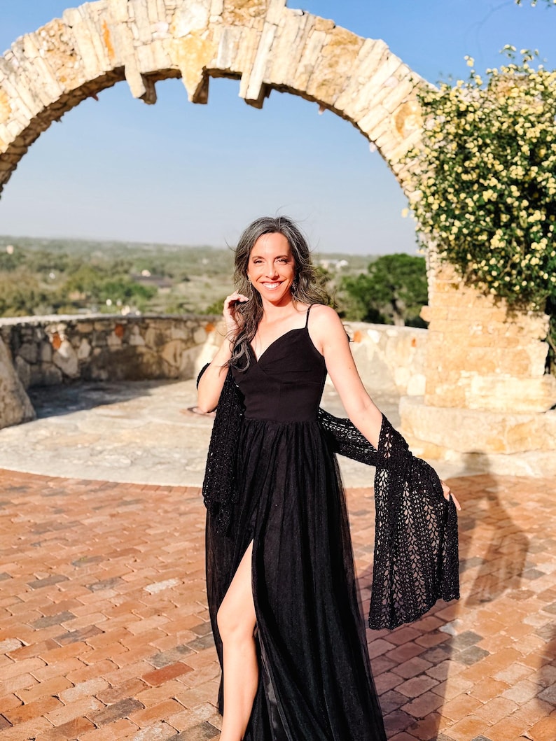 May include: A floor-length black dress with a V-neckline, thin spaghetti straps, and a high side slit, paired with a black crocheted shawl featuring an openwork pattern. The dress has a fitted bodice and a flowing, layered tulle skirt.