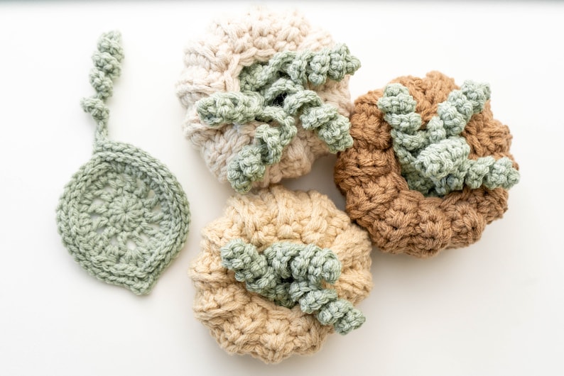 Three crochet pumpkins in cream, beige and tan with curly green stems next to a sage leaf coaster