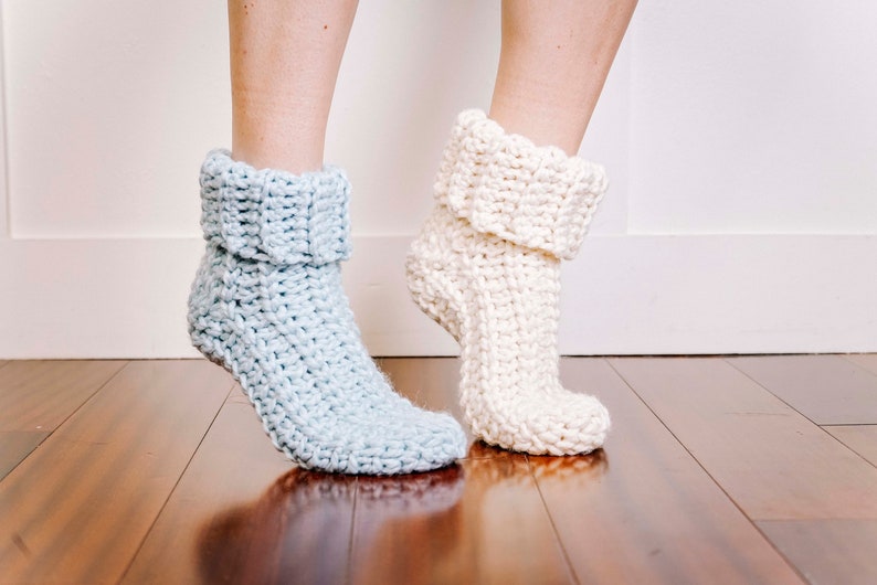 May include: A pair of crocheted slipper socks, one in light blue and one in white. The socks are folded down at the top and have a textured stitch pattern.