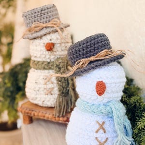 May include: Two handmade crochet snowmen. One snowman has a gray hat, orange nose, and green scarf. The other snowman has a gray hat, orange nose, and blue scarf. Both snowmen have brown stitched details and are on a wooden surface.