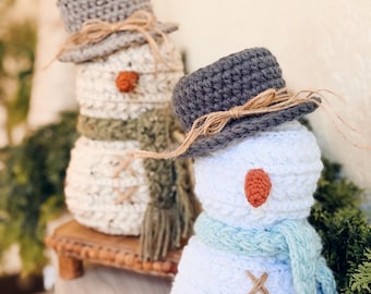 Könnte beinhalten: Zwei handgefertigte Schneemänner aus Häkelarbeit. Einer hat einen grauen Hut, eine orangefarbene Nase und einen grünen Schal. Der andere hat einen grauen Hut, eine orangefarbene Nase und einen blauen Schal. Beide haben braune Nahtdetails und stehen auf einer Holzoberfläche.