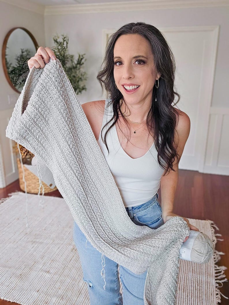 A woman displays a light gray crocheted piece of fabric, standing in a room adorned with a rug and decorative plant, offering it as a charming handmade gift.