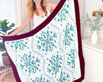 May include: A white crochet blanket with a teal geometric pattern and a burgundy border. The blanket is being held up by a person.