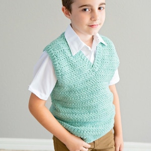 May include: A mint green, hand-crocheted vest with a V-neck, worn over a white collared shirt. The vest is paired with brown trousers. The outfit presents a classic, preppy style, suitable for children's clothing.