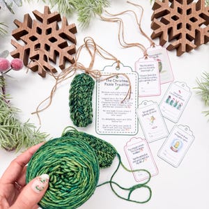 May include: A green crocheted pickle ornament with a twine hanger, a printed card titled The Christmas Pickle Tradition, and several small gift tags with illustrations and text. A ball of variegated green yarn is held in the foreground.