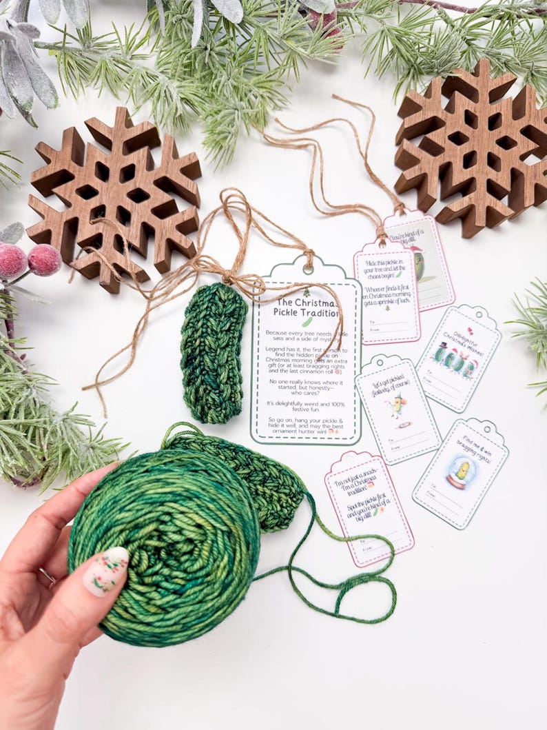May include: A green crocheted pickle ornament with a twine hanger, a printed card titled The Christmas Pickle Tradition, and several small gift tags with illustrations and text. A ball of variegated green yarn is held in the foreground.
