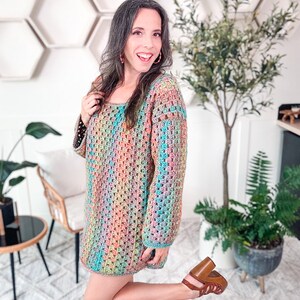 A woman stands indoors, smiling and wearing a colorful crocheted dress and brown heels. She poses with one hand on her hip and the other touching her hair, accentuating her unique jewelry. Plants and hexagonal shelves create a charming backdrop.