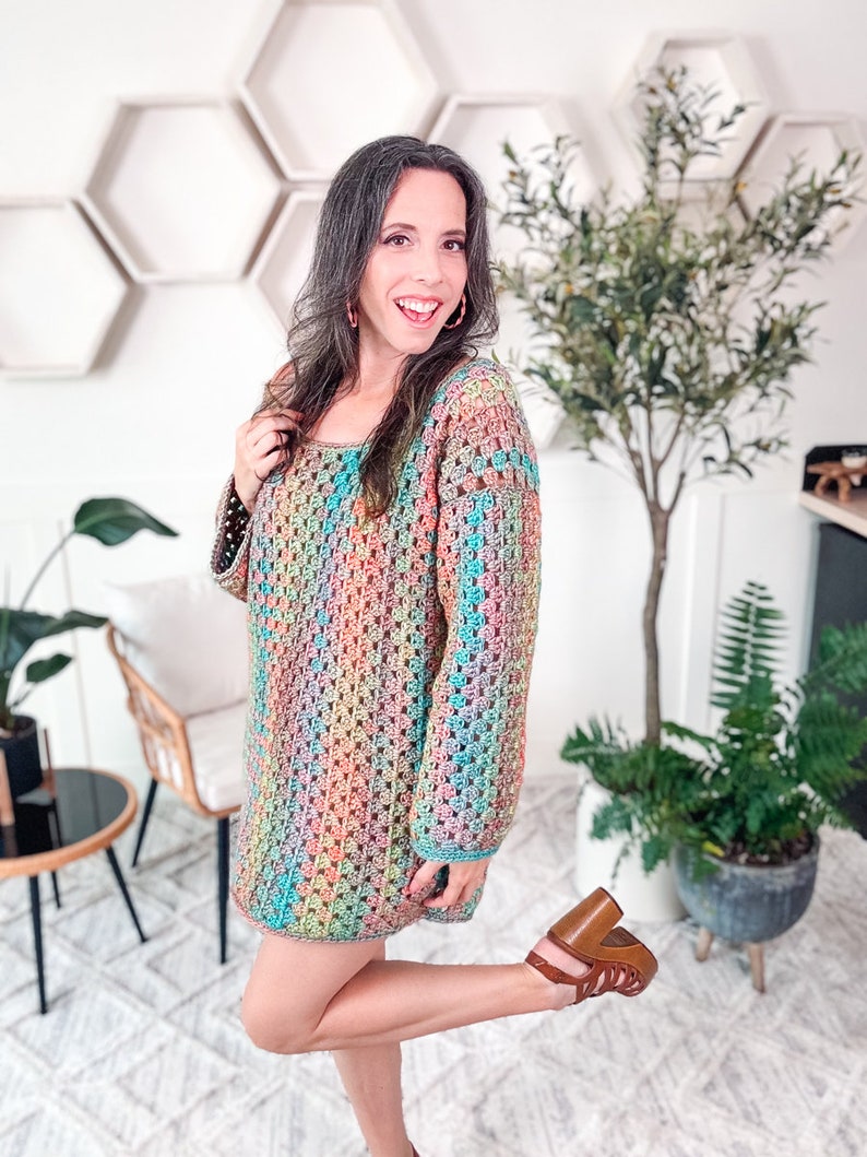 A woman stands indoors, smiling and wearing a colorful crocheted dress and brown heels. She poses with one hand on her hip and the other touching her hair, accentuating her unique jewelry. Plants and hexagonal shelves create a charming backdrop.