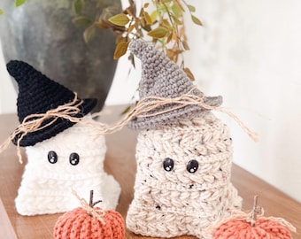 May include: Two crocheted ghost figures, one white and one speckled beige, each wearing a small pointed hat in black or grey with twine accents. Two small, ribbed, orange crocheted pumpkins sit in front of the ghosts, each topped with a twig stem.