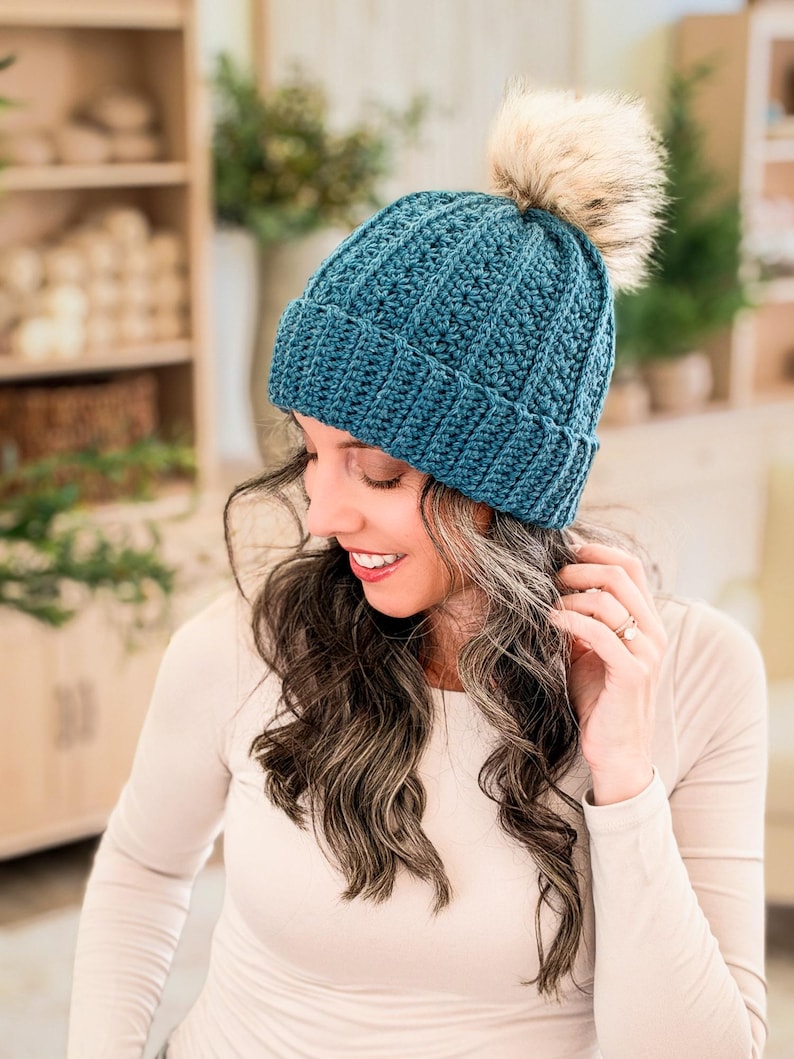 Petal Loop Beanie crochet pattern: teal crochet beanie with ribbed brim and faux fur pom pom worn by a smiling woman looking down