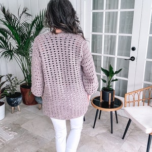 Woman standing on a patio wearing a loose-crochet sweater and white pants, facing away from the camera toward a door flanked by plants, showcasing her unique jewellery.