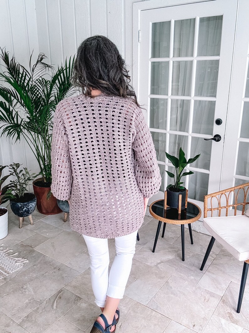 Woman standing on a patio wearing a loose-crochet sweater and white pants, facing away from the camera toward a door flanked by plants, showcasing her unique jewellery.