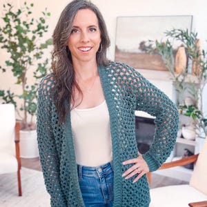 May include: A teal green crochet cardigan with an open weave design. The long-sleeved sweater has a ribbed hem and cuffs. The cardigan is worn over a white tank top and blue jeans.