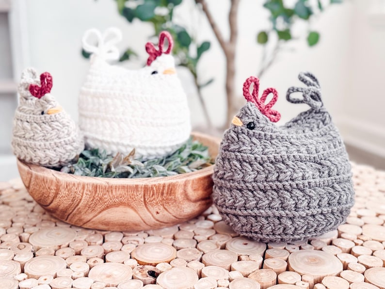 Gray, white, and oatmeal crochet chickens grouped around a wooden bowl on a wood slice tabletop