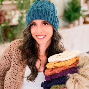 Woman in a teal crochet beanie and tan cardigan holds a stack of beanies in rainbow colors