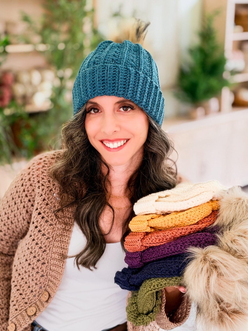 Woman in a teal crochet beanie and tan cardigan holds a stack of beanies in rainbow colors