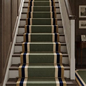 Könnte beinhalten: Eine Treppe mit einem Läufer, der ein zentrales olivgrünes Feld aufweist, flankiert von cremefarbenen und marineblauen Streifen. Der Läufer ist mit Metallstangen an jeder Stufe befestigt, vor weißen Setzstufen und dunklen Holztrittstufen.