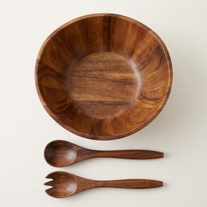 Könnte beinhalten: Eine runde Salatschüssel aus Holz mit natürlicher brauner Maserung, begleitet von einem passenden hölzernen Servierlöffel und einer hölzernen Serviergabel mit drei Zinken, die zusammen auf einer hellen, einfarbigen Oberfläche liegen.