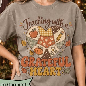 May include: A sandstone-coloured t-shirt featuring a graphic of a heart-shaped quilt pattern with an apple, pencil, pumpkin, and flower. The text reads Teaching with a GRATEFUL HEART in brown and orange lettering, surrounded by small autumn leaves.