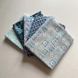 May include: A stack of five folded fabric squares featuring various patterns. The top piece is grey with a blue and pink floral grid. Other pieces include a blue scalloped pattern, a small pink floral print, and two dark blue floral designs.