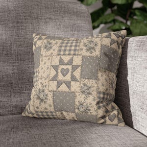 May include: A square throw pillow featuring a patchwork design in shades of gray and cream. The pattern includes floral prints, gingham checks, polka dots, and a central star motif containing a heart shape. The fabric has a quilted texture.
