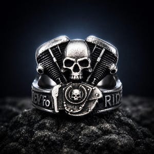 Könnte beinhalten: Silberfarbener Ring mit Totenkopf-Design in der Mitte, flankiert von Motorkomponenten. Der Ring hat die Gravur "NEVER TO RIDE". Der Ring ist vor einem dunklen Hintergrund platziert.
