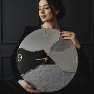 May include: A round wall clock featuring a segmented design in shades of black, dark gray, and light gray. The surface has varied textures, including smooth and rough sections separated by gold-colored lines. Gold numerals 3, 6, 9, and 12 are present.