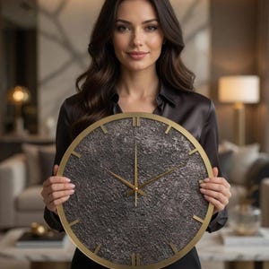 May include: A large, round wall clock with a textured dark grey face and gold-coloured hands and frame. The clock features Roman numeral hour markers. The clock is being held by a person.