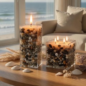 Puede incluir: Dos portavelas cuadrados de cristal rellenos de piedras de río lisas y multicolores. La vela más alta tiene una mecha encendida, mientras que la más corta y ancha tiene cuatro. Ambas velas contienen cera de color claro y emiten un brillo.