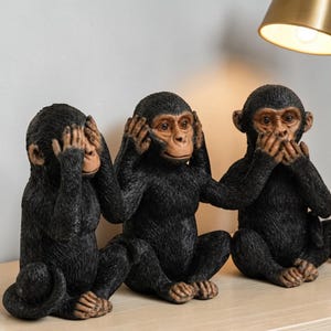May include: Three black monkey ornaments in the classic "See no evil, hear no evil, speak no evil" pose. The monkeys have brown faces and hands, and are sitting down. The ornaments are on a light-coloured surface.