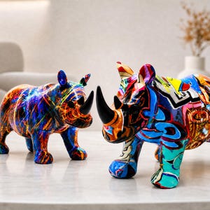 Peut inclure: Deux figurines de rhinocéros aux motifs multicolores vibrants. Le rhinocéros de gauche présente des traînées tourbillonnantes orange, bleues et jaunes, tandis que celui de droite arbore des formes graffiti audacieuses en bleu, rouge, jaune et noir. Les deux ont des cornes noires unies.