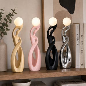 May include: Four abstract sculptural table lamps featuring a smooth, curved base in matte yellow, matte pink, matte black, and polished silver. Each lamp is topped with a spherical white glass light bulb, creating a balanced and modern lighting display.