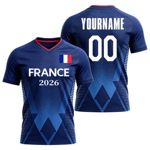 K&ouml;nnte beinhalten: Ein dunkelblaues Kurzarmtrikot mit V-Ausschnitt und rotem Innenbesatz. Die Vorderseite zeigt die franz&ouml;sische Flagge, das Wort FRANCE und das Jahr 2026 in Wei&szlig;. Die R&uuml;ckseite zeigt den Text YOURNAME und die Nummer 00 in Wei&szlig; auf einem geometrischen Muster.