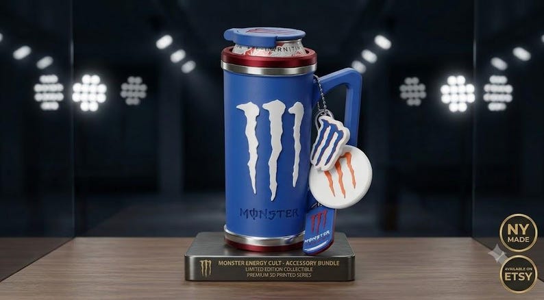 K&ouml;nnte beinhalten: Ein blauer zylindrischer Dosenhalter mit wei&szlig;em Monster Energy Logo und Schriftzug. Er verf&uuml;gt &uuml;ber einen Griff, einen roten Rand und drei h&auml;ngende Anh&auml;nger. Die Basis ist eine rechteckige Plakette mit der Aufschrift Monster Energy Cult.