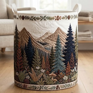 May include: A cylindrical fabric laundry basket featuring a quilted white background with an embroidered landscape of brown mountains, dark green and orange pine trees, ferns, and mushrooms. The basket has two white fabric handles at the top.
