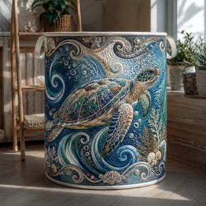 May include: A cylindrical fabric laundry hamper featuring a detailed quilted design of a sea turtle swimming through blue ocean waves, coral, and seashells. The hamper has two white side handles and a white top rim, showcasing intricate textures.