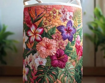 May include: A cylindrical fabric laundry hamper featuring a quilted texture and a vibrant tropical print. The design displays various flowers including hibiscus, orchids, and plumeria, alongside green monstera leaves on an orange background.