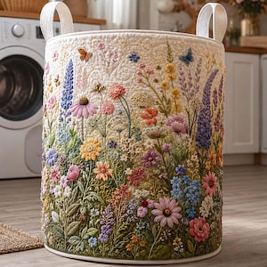 Peut inclure: Un panier à linge cylindrique en tissu matelassé de couleur crème, orné d'un motif complexe de broderies en relief représentant des fleurs sauvages colorées, du feuillage vert, des papillons et une petite abeille, avec deux anses blanches.